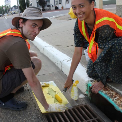Two volunteers kneel beside a storm drain where they use a stencil to paint a yellow fish